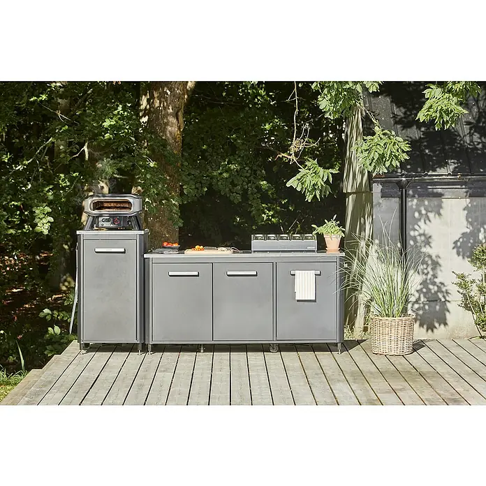 Cozze Pizzaofen Premium 13" - Grillfläche (B x T): 34 x 34 cm, 6 kW - Außenküche mit Pizzaofen und Kochfeld auf einer Holzterrasse, umgeben von Grünpflanzen. Die graue Küche bietet Stauraum.