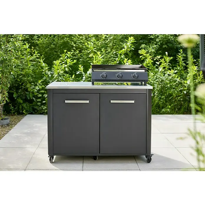 Cozze Plancha-Grill G 700Grillfläche (B x T): 66,5 x 35 cm, Anzahl Brenner: 3 Stk., Schwarz - Schwarzer, mobiler Outdoor-Küchenschrank mit Grillplatte auf einer hellen Steinterrasse vor üppigem Grün. Praktische Grilllösung für den Garten.