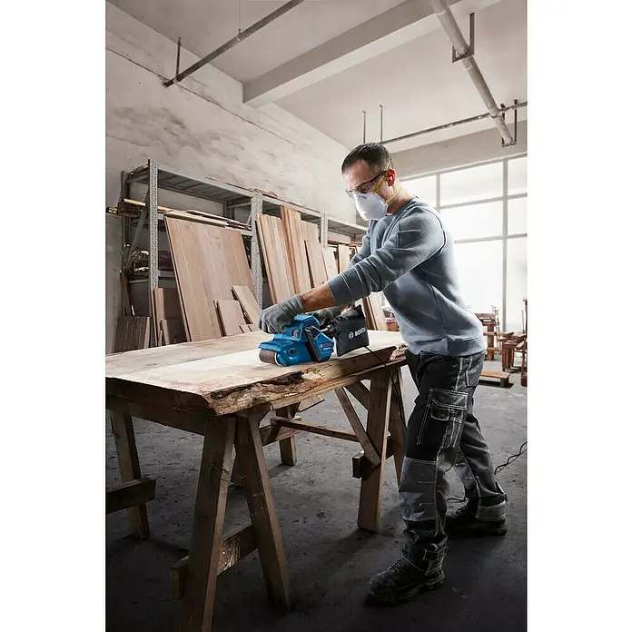 Bosch Professional Elektro-Bandschleifmaschine GBS 750 - 850 W - Mann schleift Holz mit einem Bosch Bandschleifer in einer Werkstatt, Schutzmaßnahmen inklusive.