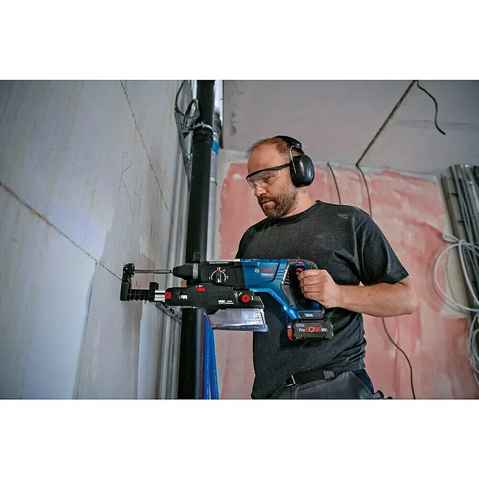 Bosch Professional AMPShare 18V Akku-Bohrhammer GBH 18V-28 D - 18 V, Ohne Akku - Ein Mann mit Schutzbrille und Kopfhörern bohrt mit einem blauen Boschhammer in eine Wand, wobei er einen Staubsaugeraufsatz verwendet.
