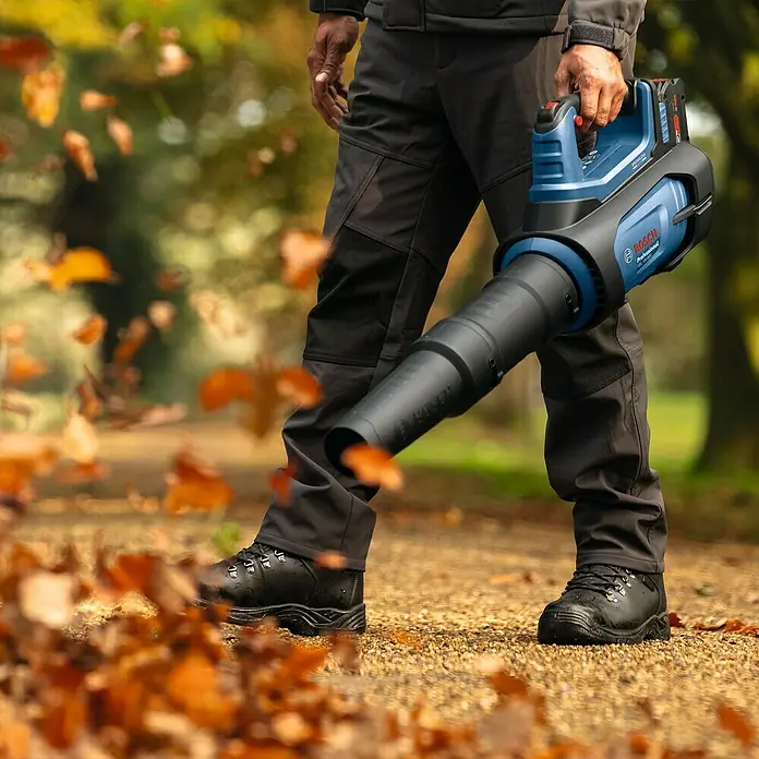 Bosch Professional AMPShare 18V Akku-Laubbläser GBL 18V-800 - 18 V, Ohne Akku, Blasgeschwindigkeit: 200 km/h, Blasleistung: 800 m³/h, Länge: 85,5 cm - Ein Mann benutzt einen Bosch Akku-Laubbläser, um Herbstlaub auf einem Weg zu entfernen. Das Gerät ist blau und schwarz und mit dem Bosch Professional Logo versehen.