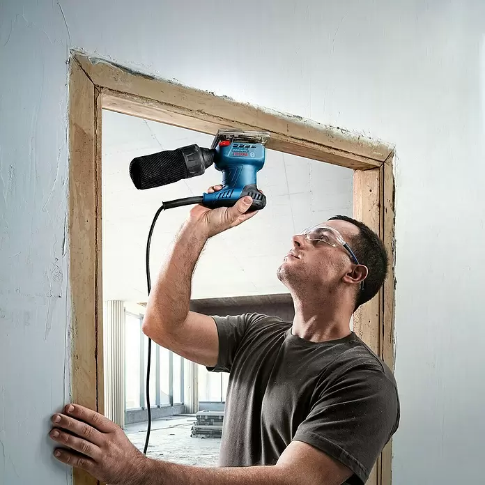 Bosch Professional Elektro-Schwingschleifer GSS 140 A - Leerlaufschwingzahl: 28.000 U/min, Durchmesser Schwingkreis: 1,5 mm - Mann benutzt blauen Bosch Schleifer mit schwarzem Staubbehälter zum Schleifen eines Holz-Türrahmens. Er trägt Schutzbrille.