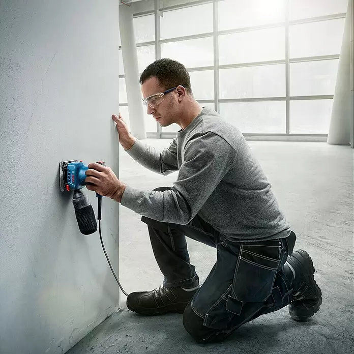 Bosch Professional Elektro-Schwingschleifer GSS 140 A - Leerlaufschwingzahl: 28.000 U/min, Durchmesser Schwingkreis: 1,5 mm - Mann nutzt ein blaues Bosch Elektrowerkzeug mit Staubbeutel zum Schleifen einer hellgrauen Wand.