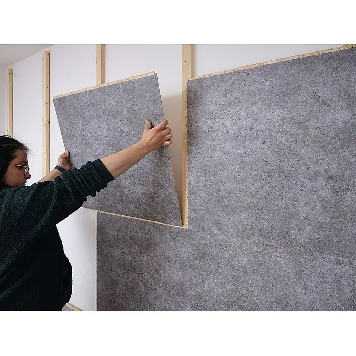 Egger Wandpaneel DecoWall - Rovigo Beton, 125 cm x 66 cm x 12 mm - Eine Person bringt eine graue Wandfliese mit Holzrahmen an einer Wand an, die ebenfalls mit Holzlatten versehen ist.
