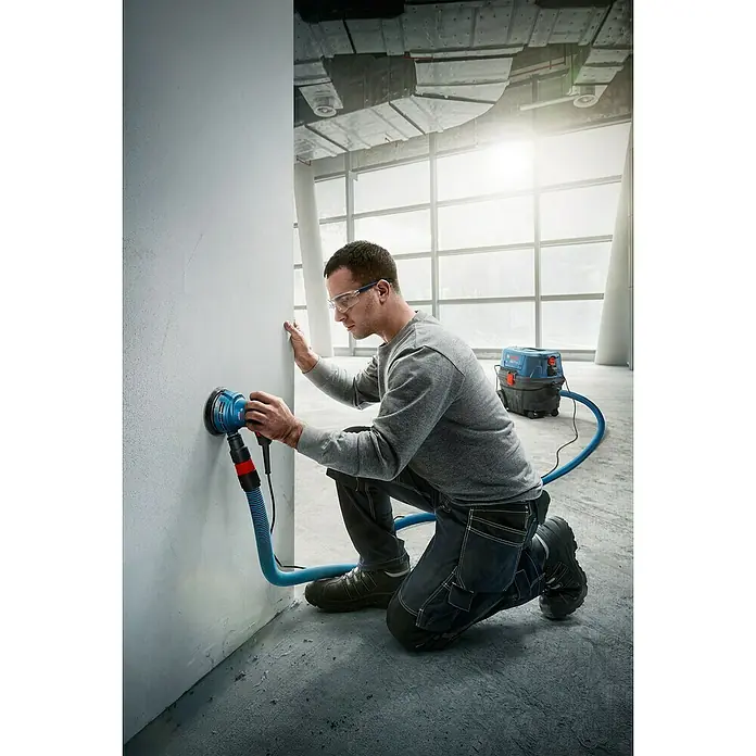 Bosch Professional Elektro-Exzenterschleifer GEX 125 - 290 W, Durchmesser Schleifteller: 125 mm - Ein Handwerker schleift eine Wand mit einem blauen Bosch Exzenterschleifer, der an einen blauen Staubsauger angeschlossen ist.