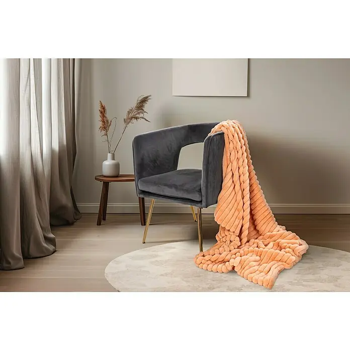 Decke Plush - Peach, 200 x 150 cm, 100 % Polyester - Nahaufnahme eines grauen Sessels mit einer orangefarbenen Strickdecke, die elegant drapiert ist, auf einem hellen, runden Teppich.