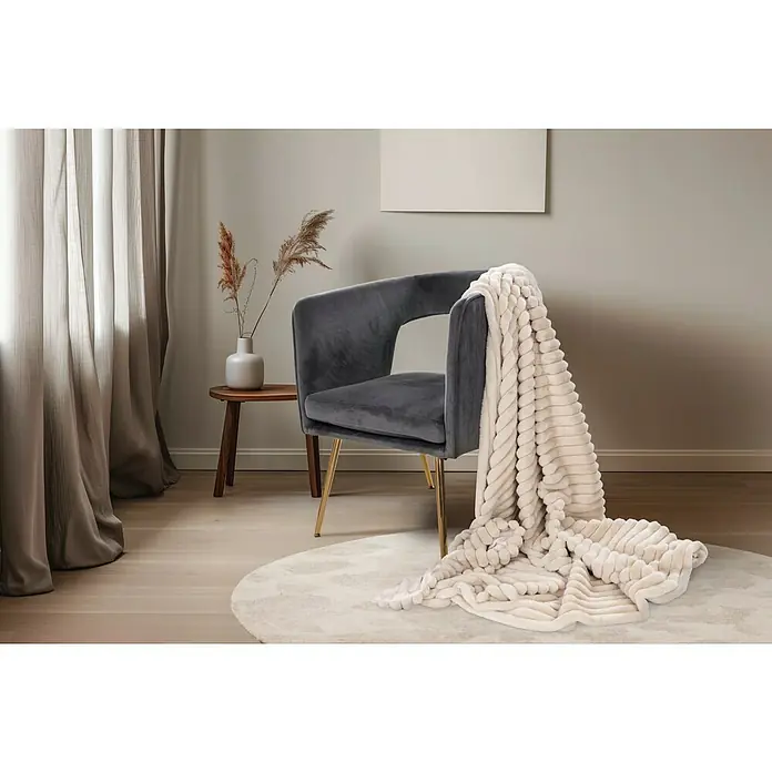 Decke Plush - Beige, 200 x 150 cm, 100 % Polyester - Ein grauer Sessel mit goldenen Beinen und einer cremefarbenen Decke darauf, auf einem runden Teppich in einem hellen Raum.