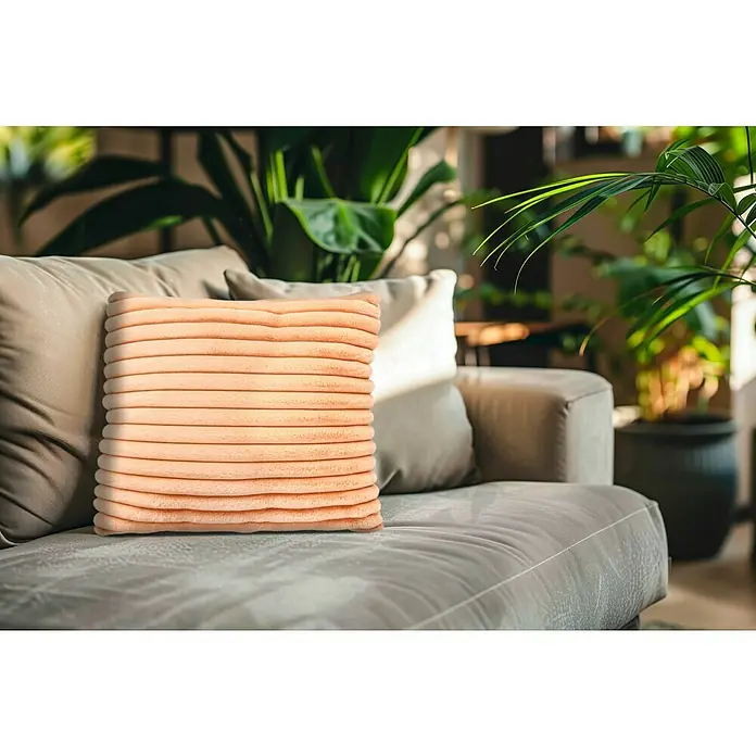 Kissen Plush - Peach, 45 x 45 cm, 100 % Polyester - Nahaufnahme eines gerippten, pfirsichfarbenen Kissens auf einem grauen Sofa, umgeben von üppigem Grün, das ein modernes, einladendes Ambiente ausstrahlt.