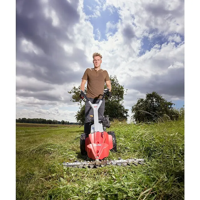AL-KO Comfort Frontbalkenmäher 875 IV -  - Ein junger Mann bedient eine rote AL-KO Wiesenmähermaschine auf einem grünen Feld, bewölkter Himmel im Hintergrund.