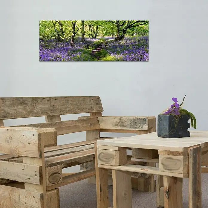 Glasbild - To Promenade, B x H: 80 x 30 cm - Detailaufnahme eines Wohnraumes mit einer Couch und einem Tisch aus Holzpaletten sowie einem gerahmten Landschaftsbild an der Wand.