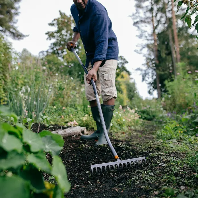 Fiskars Ergonomic Gartenrechen - Material Gerätekopf: Borstahl, 1 Stk. - Ein Mann harkt mit einem Fiskars Rechen einen Gartenweg. Der Rechen ist silbern und hat einen orangen Akzent am Kopf.