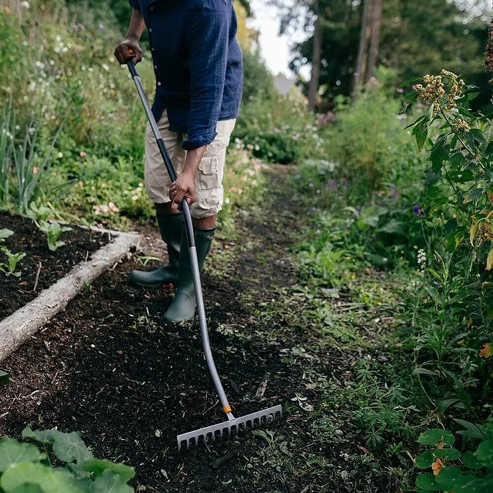 Fiskars Ergonomic Gartenrechen - Material Gerätekopf: Borstahl, 1 Stk. - Eine Person benutzt einen Fiskars Rechen im Garten, um den Boden vorzubereiten. Der Rechen ist ideal für die Gartenarbeit.
