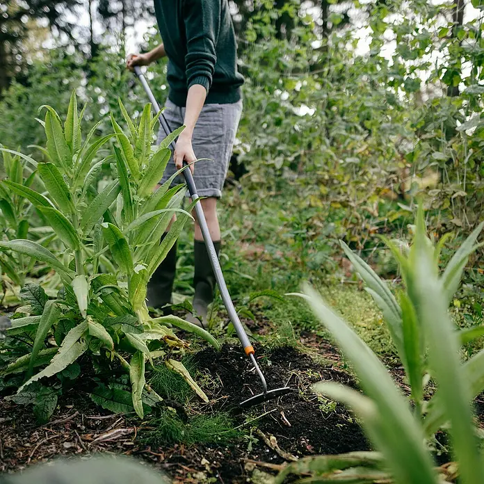 Fiskars Ergonomic Unkrauthacke - Länge: 154 cm, Material Gerätekopf: Borstahl - Die Aufnahme zeigt eine Person, die mit einem Fiskars Rechen im Garten arbeitet, um den Boden aufzulockern.