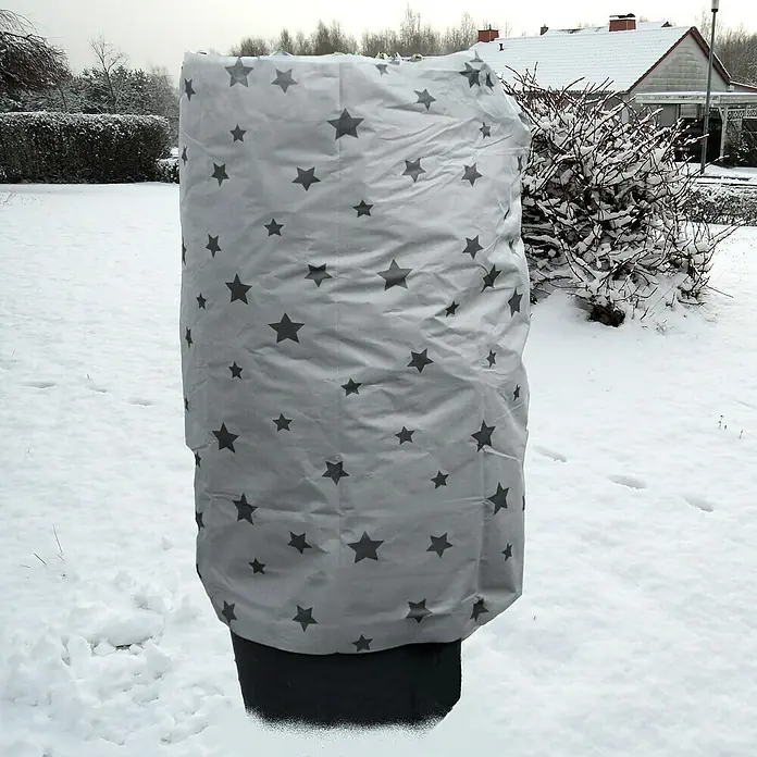 Winterschutzvlies - 5 x 1,5 m, Grau, Grammatur: 50 g/m² - Nahaufnahme einer Pflanze, die mit einer Schutzhülle mit Sternmuster vor der Kälte geschützt ist, im verschneiten Garten.