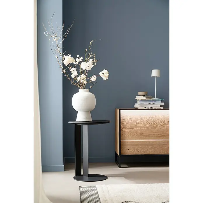 SCHÖNER WOHNEN-Kollektion Sockelleiste Modern 5 - 200 cm x 58 mm x 13 mm - Elegantes Interieur mit einer stilvollen weißen Vase auf einem schwarzen Beistelltisch, ergänzt durch ein Sideboard mit Büchern und einer Lampe.