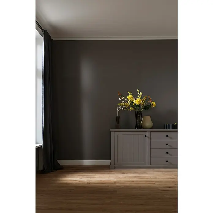 SCHÖNER WOHNEN-Kollektion Sockelleiste Modern 6 - 200 cm x 110 mm x 15 mm, Hochverdichtetes extrudiertes Polymer (HDPS) - Grauer Holzschrank mit Schubladen und Türen unter einem Blumenstrauß und einer Kerze, vor einer grauen Wand und Parkettboden.