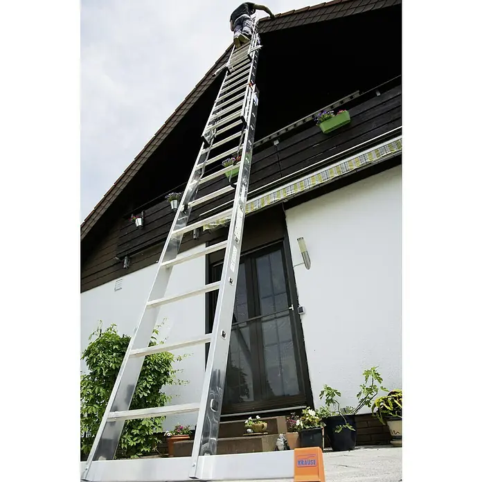 Krause Monto Mehrzweckleiter Tribilo - Arbeitshöhe: 9,3 m, Anzahl Tritte: 36 Stk., Belastbarkeit: 150 kg, Aluminium - Eine lange, ausziehbare Leiter steht an einem Haus, auf der ein Mann steht, der am Dach arbeitet. Die Leiter ist von der Marke Krause.