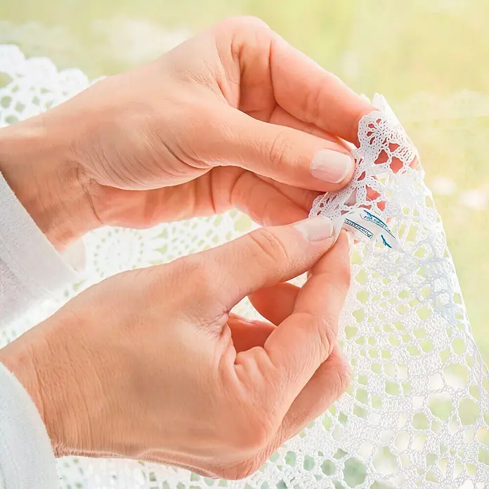 Tesa Powerstrips Cinta engomada - Transparente - Una persona sostiene un tapete de ganchillo blanco, examinando la etiqueta. El intrincado diseño del tapete es claramente visible.