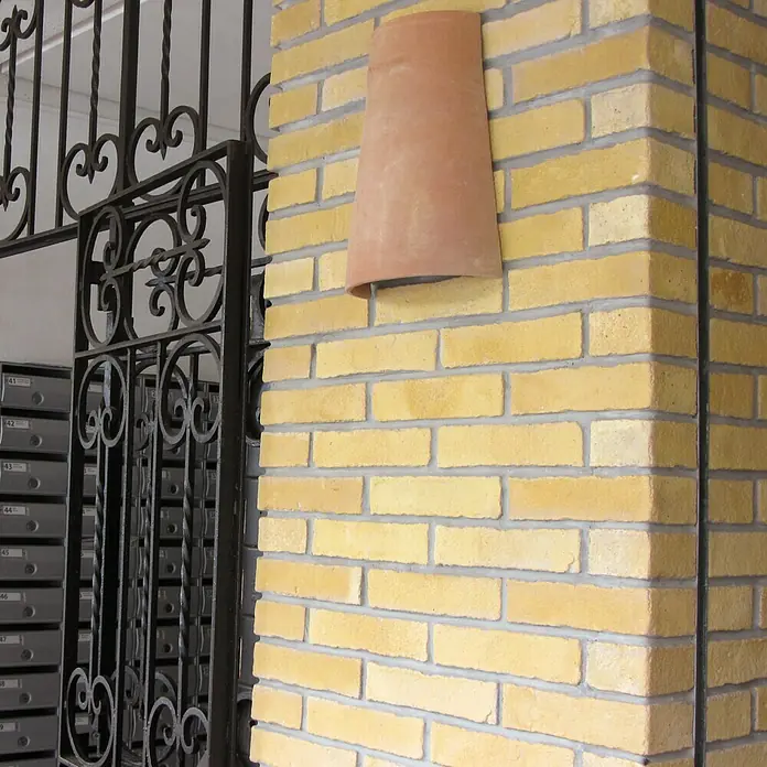 Esquina exterior Mediterráneo Barro - 24 x 12 x 5,5 cm - Primer plano de un elemento de pared de terracota en forma de tubo fijado a una pared de ladrillo amarillo, que ofrece una estética rústica.
