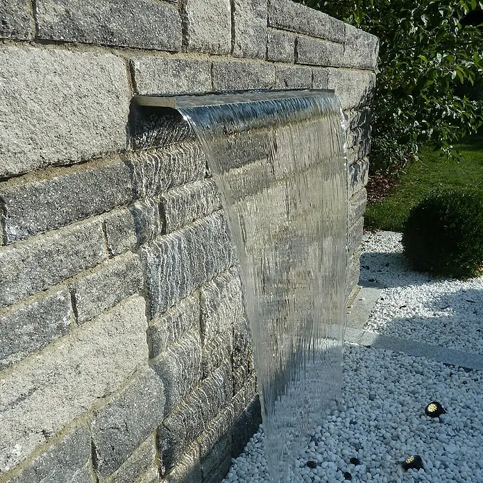 Heissner Einbau-Wasserfall W483 - 30 x 18 cm, Edelstahl - Nahaufnahme eines modernen Wasserfalls an einer grauen Steinmauer mit weißen Kieselsteinen.