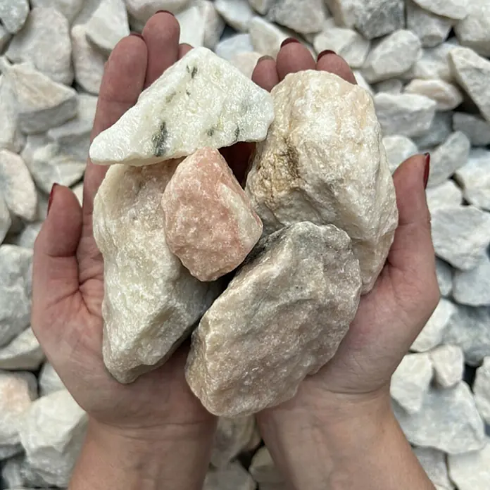 Min2C Bruchsteine - Rose Sölker, Körnung: 30 mm - 60 mm, 25 kg - Nahaufnahme von rosa und weißen Marmorkieseln in den Händen, ideal für Gartenwege und dekorative Zwecke.