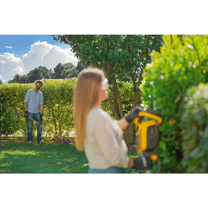 Stiga 500er Serie Akku-Rasentrimmer GT 500e - 48 V, 2 Ah, Li-Ionen, Ohne Akku, Schnittbreite Faden: 30 cm - Eine Frau trimmt die Hecke mit einer gelben Heckenschere, während ein Mann im Hintergrund mit einem Rasentrimmer arbeitet.