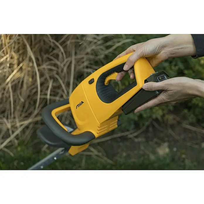 Stiga 100er Serie Akku-Heckenschere HT 100e Set - 20 V, Li-Ionen, 1 Akku, Schnittlänge: 50 cm - Nahaufnahme einer gelben STIGA-Heckenschere mit eingesetztem Akku, gehalten von Händen.
