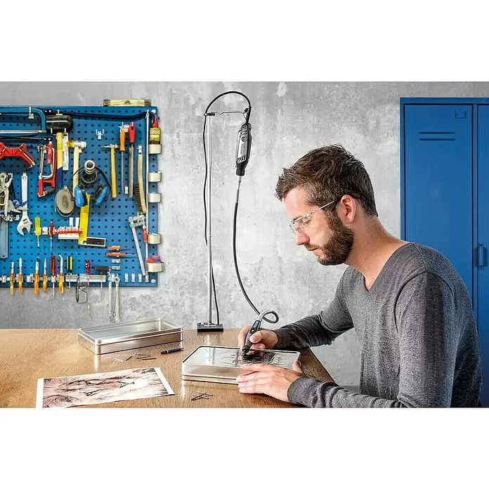 Dremel Soporte para herramientas 2222 - Apto para: Multi-Herramientas - Un hombre graba un diseño en una lata de metal con una herramienta rotatoria gris y negra suspendida de un soporte.