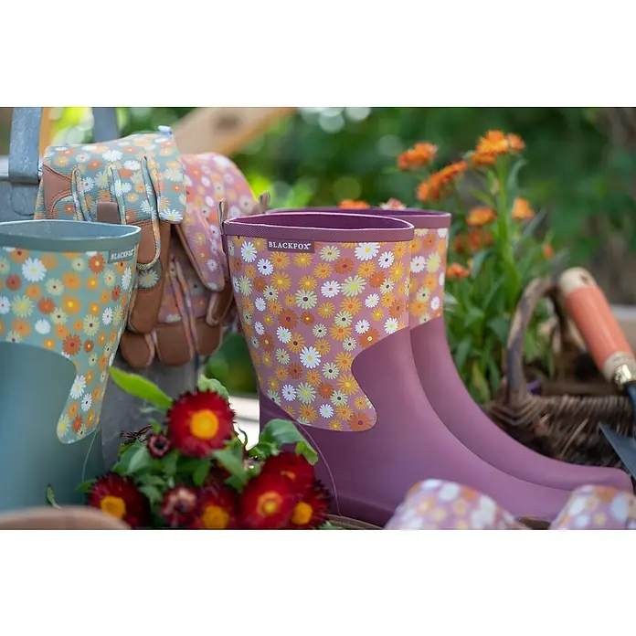 Black Fox Gummistiefel Lizia - Schuhgröße: 40, Rosa - Gartenszene mit Blumenstiefeln und Gartenhandschuhen mit Blumenmuster, die das Bild mit botanischem Charme erfüllen.