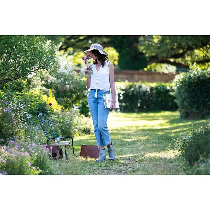 Black Fox Gummistiefel Bella - Schuhgröße: 38, Grau - Eine Frau mit Hut und Blumen steht in einem üppigen Garten mit einem Notizbuch in der Hand. Die Szene strahlt Ruhe und Inspiration aus.