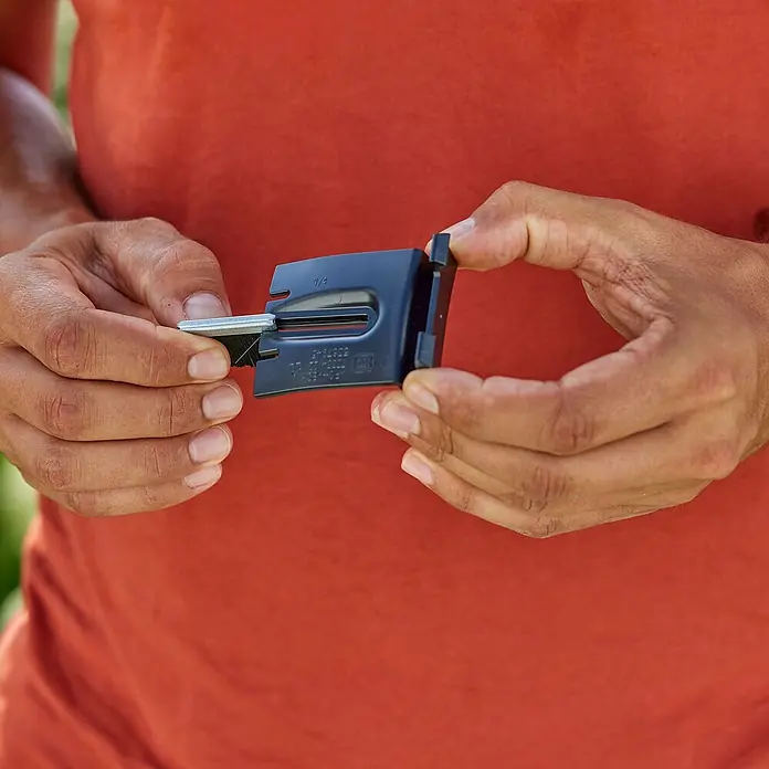 Gardena Bürsten-Set für Rasenmäher - Gardena Mähroboter smart SILENO pro, smart SILENO max und smart SILENO free - Eine Nahaufnahme einer Person, die ein kleines, dunkles Werkzeug mit einer Metallklinge in der Hand hält. Das Werkzeug wird vor einem roten Hemd gehalten.