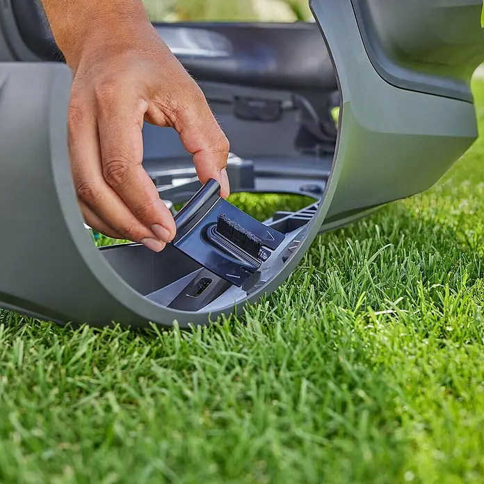 Gardena Bürsten-Set für Rasenmäher - Gardena Mähroboter smart SILENO pro, smart SILENO max und smart SILENO free - Eine Hand befestigt eine schwarze Bürste an einem Rasenmähroboter auf einem grünen Rasen.