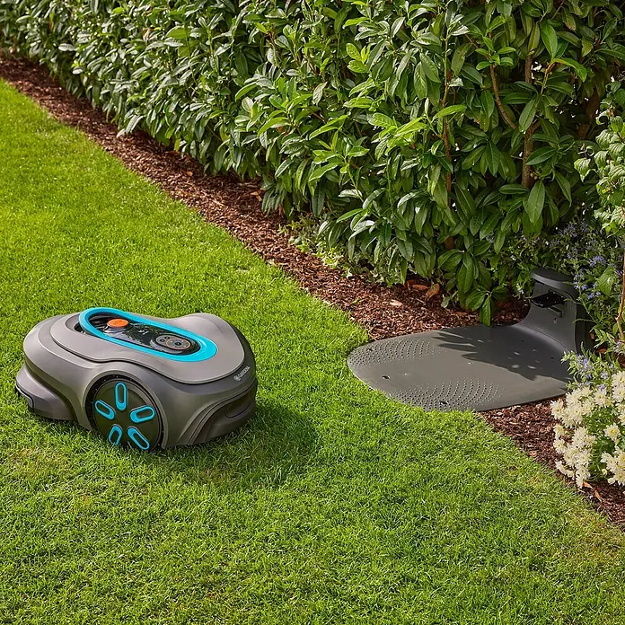 Gardena Mähroboter smart SILENO pro 600 m² - 600 m², Schnittbreite: 16 cm, Induktionsgestützte Navigation, Begrenzungskabel - Ein Gardena Mähroboter neben seiner Ladestation auf einem grünen Rasen vor einer Hecke.