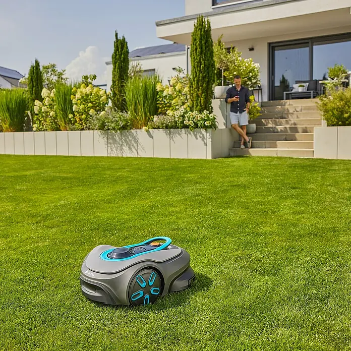 Gardena Mähroboter smart SILENO free - 600 m², Schnittbreite: 22 cm, Satellitengestützte Navigation, GPS - Ein Gardena Mähroboter auf einem grünen Rasen mit einem Mann im Hintergrund, der ein Handy benutzt.