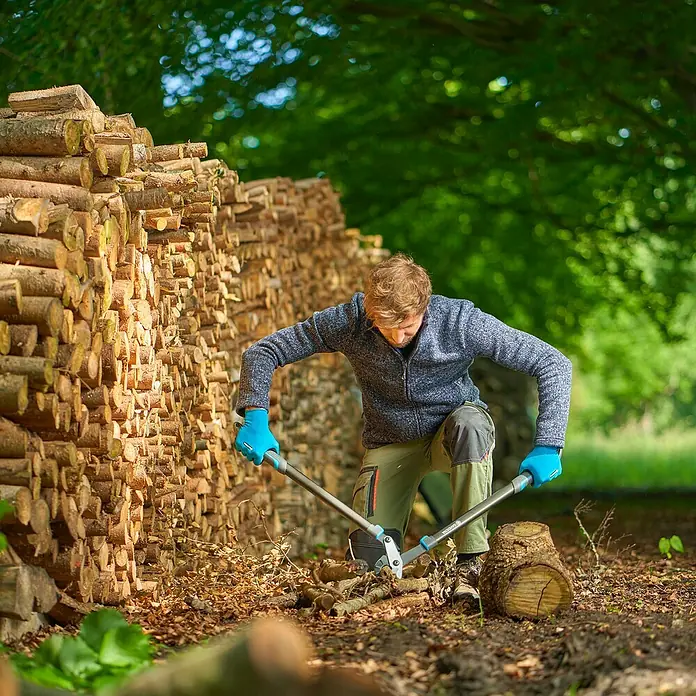 Gardena EnergyCut Astschere Pro L - Schneidsystem Gartenschere: Amboss, Max. Aststärke: 50 mm - Ein Mann spaltet Holz mit einer Gardena Astschere vor einem Holzstapel, umgeben von Bäumen. Ideal für Gartenarbeiten.