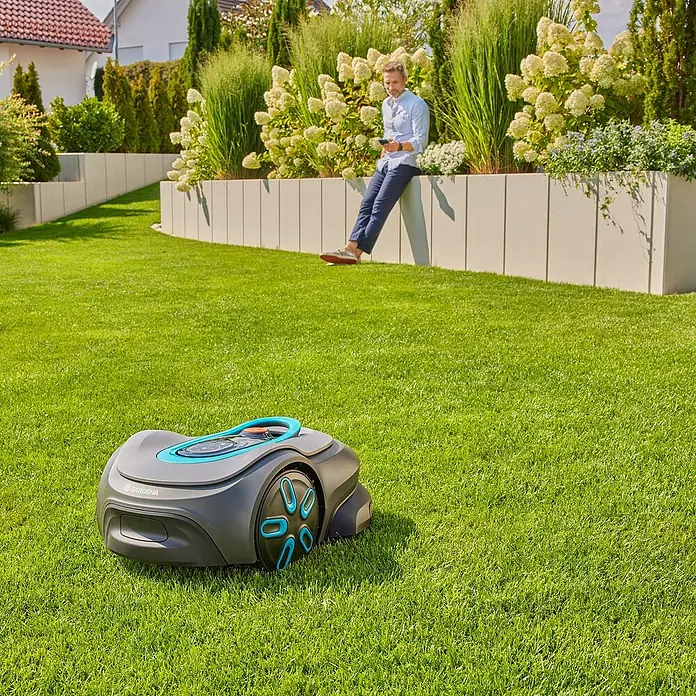 Gardena Mähroboter smart SILENO max - 1.500 m², Schnittbreite: 22 cm, Induktionsgestützte Navigation, Begrenzungskabel - Ein Gardena Mähroboter steht auf einem gepflegten Rasen, während ein Mann sich an einer Mauer mit Blumenkästen entspannt.