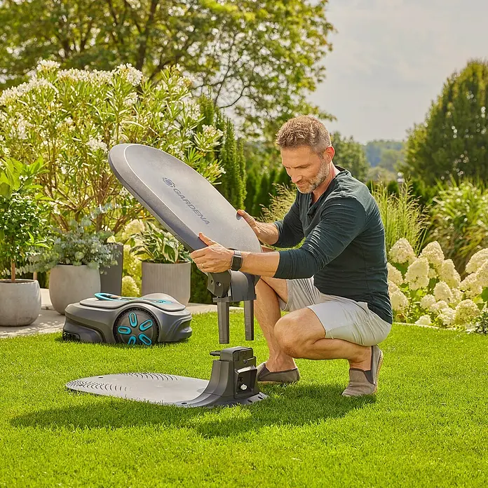 Gardena Mähroboter-GarageGardena Mähroboter smart SILENO pro / max / free - Ein Mann montiert eine Gardena Ladestation für Rasenmähroboter im Garten. Der Rasenmäher steht daneben bereit für den Einsatz.