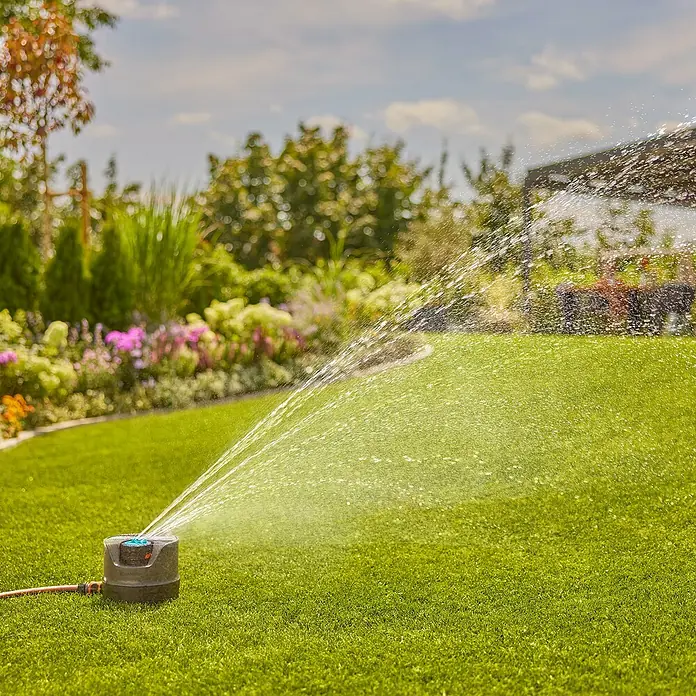 Gardena Vielflächenregner AquaPrecise solarbetrieben - Max. Regnerfläche: 320 m², Wurfweite: 2 m - 10 m - Automatisch rotierender Rasensprenger im Garten bei der Bewässerung des grünen Rasens und der Pflanzen im Hintergrund.