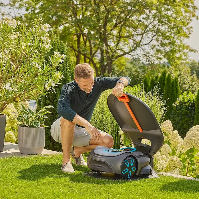 Gardena Mähroboter-GarageGardena Mähroboter smart SILENO pro / max / free - Ein Mann hockt neben einem Gardena Mähroboter mit geöffneter Klappe, umgeben von grüner Rasenfläche und Pflanzen.