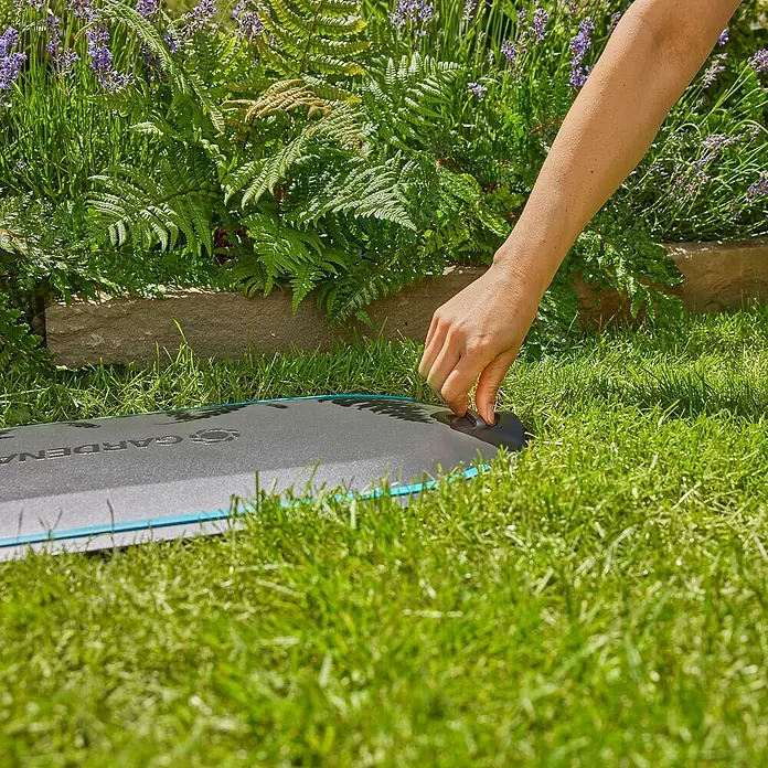 Gardena Schlauchbox Undercover RollUp - Schlauchlänge: 20 m - Nahaufnahme einer Hand, die an einem Gardena-Produkt im Gras hantiert, mit üppigem Grün im Hintergrund.