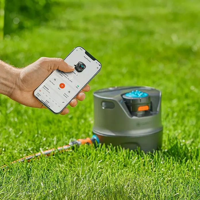 Gardena Vielflächenregner AquaPrecise solarbetrieben - Max. Regnerfläche: 320 m², Wurfweite: 2 m - 10 m - Das Bild zeigt einen smarten Gardena AquaPrecise Bewässerungscomputer, der per Smartphone-App gesteuert wird, um die Gartenbewässerung zu automatisieren.