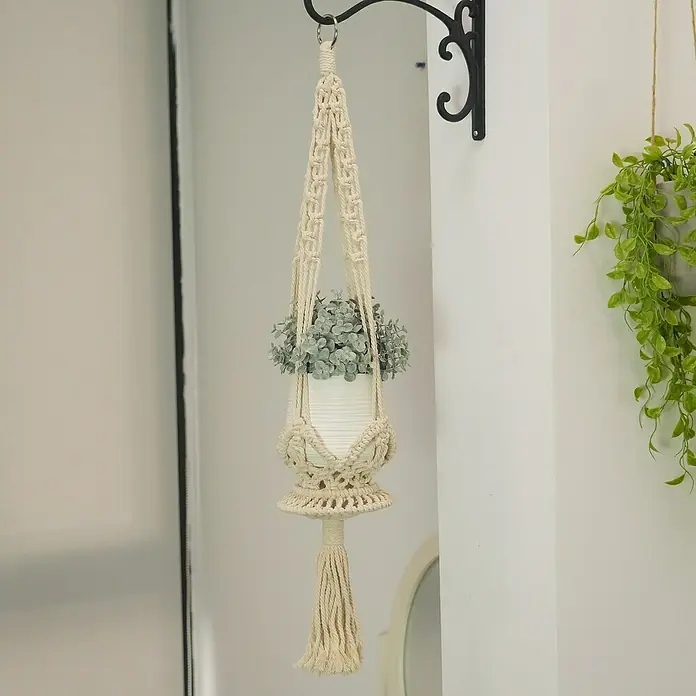 Blumenampel Makramee - Ø x H: 4 x 80 cm, Beige - Eine Makramee-Pflanzenaufhängung mit einem Topf mit Pflanze hängt an der Wand, was einen Hauch von Boho-Chic verleiht.