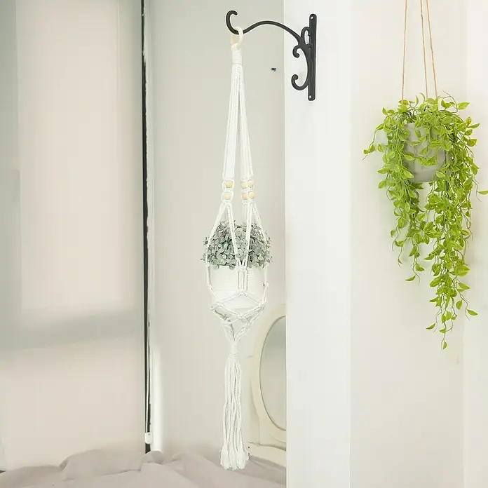 Blumenampel Makramee - Ø x H: 4 x 110 cm, Weiß - Eine Nahaufnahme eines Makramee-Pflanzenaufhängers mit einer Pflanze darin, der an einem verzierten schwarzen Haken an einer weißen Wand hängt.