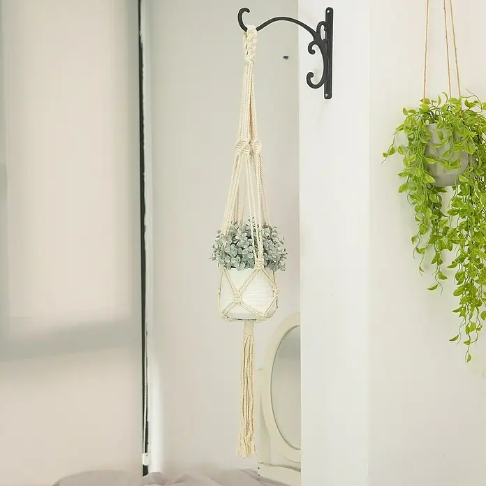 Blumenampel Makramee - Ø x H: 4 x 105 cm, Beige - Die Nahaufnahme zeigt einen stilvollen, hängenden Makramee-Blumentopf mit einer eleganten weißen Topfblume an einer schwarzen Wandhalterung.