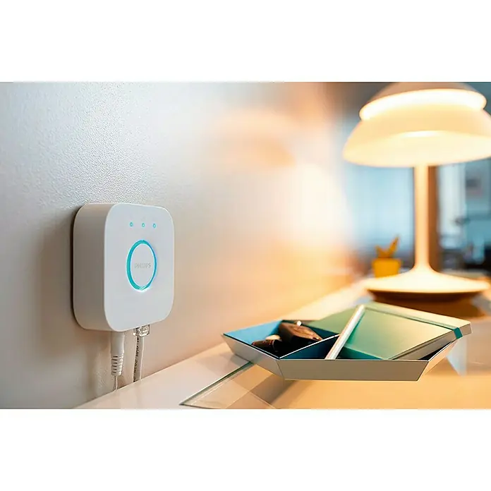 Philips Hue Steuerzentrale Bridge - Passend für: Philips Hue System, L x B x H: 2,6 x 8 x 8 cm - Nahaufnahme eines Philips Hue Bridge Smart Home Hub an einer Wand, verbunden mit Kabeln, neben einem Nachttisch mit Lampe.
