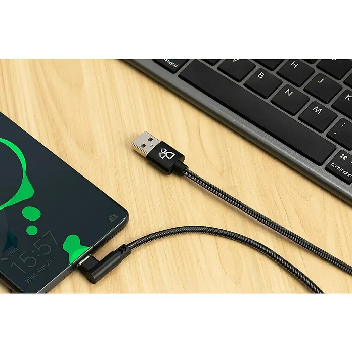 BAUHAUS USB-Ladekabel Winkel - Schwarz, 1 m, USB A-Stecker, USB C-Stecker - Ein Smartphone wird mit einem robusten USB-Kabel mit einem markanten Logo am Stecker aufgeladen, neben einer Computertastatur.