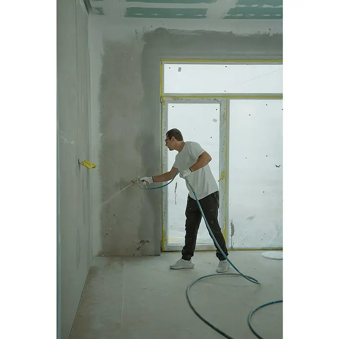 Graco Magnum Sistema de pulverización de pintura A100 Proplus - Caudal: 1,8 l/min, Velocidad de trabajo: 18 m²/min - Un hombre en construcción está trabajando en una pared con un rociador de pintura para repintar la pared mientras que una tubería azul está conectada.