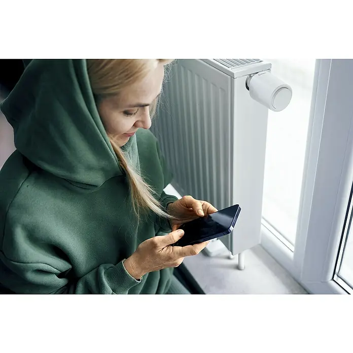 Ledvance Smart+ WiFi Heizkörper-Thermostat 1 Stück - M30 x 1,5 mm, 1 Stk. - Frau mit Kapuzenpulli, die neben einem Heizkörper mit Thermostatregler auf ihr Smartphone schaut.