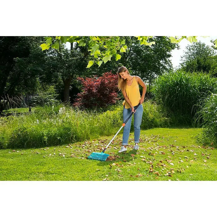 Gardena Combisystem Verlängerungsstiel - 0,9 m - 1,45 m - Frau kehrt Blätter mit einem blauen Gardena-Laubbesen auf einer Rasenfläche mit Bäumen.
