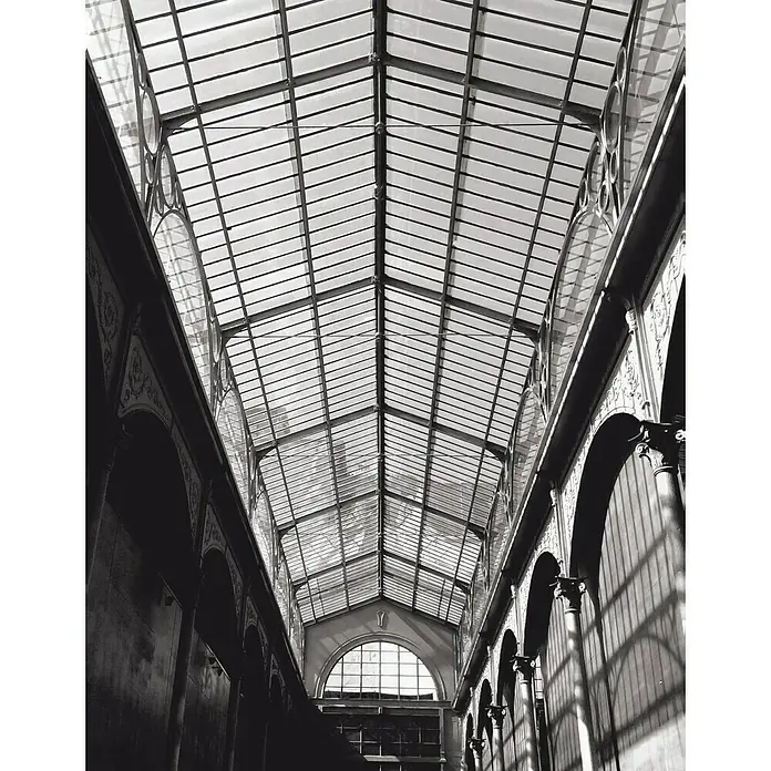 Rasch Fototapete Digitaldruck Industriehalle - B x H: 300 x 233 cm, Vlies - Schwarz-Weiß-Foto einer eleganten Passage mit Glasdach, das filigrane Metallstrukturen und sanftes Lichtspiel zeigt.\n
